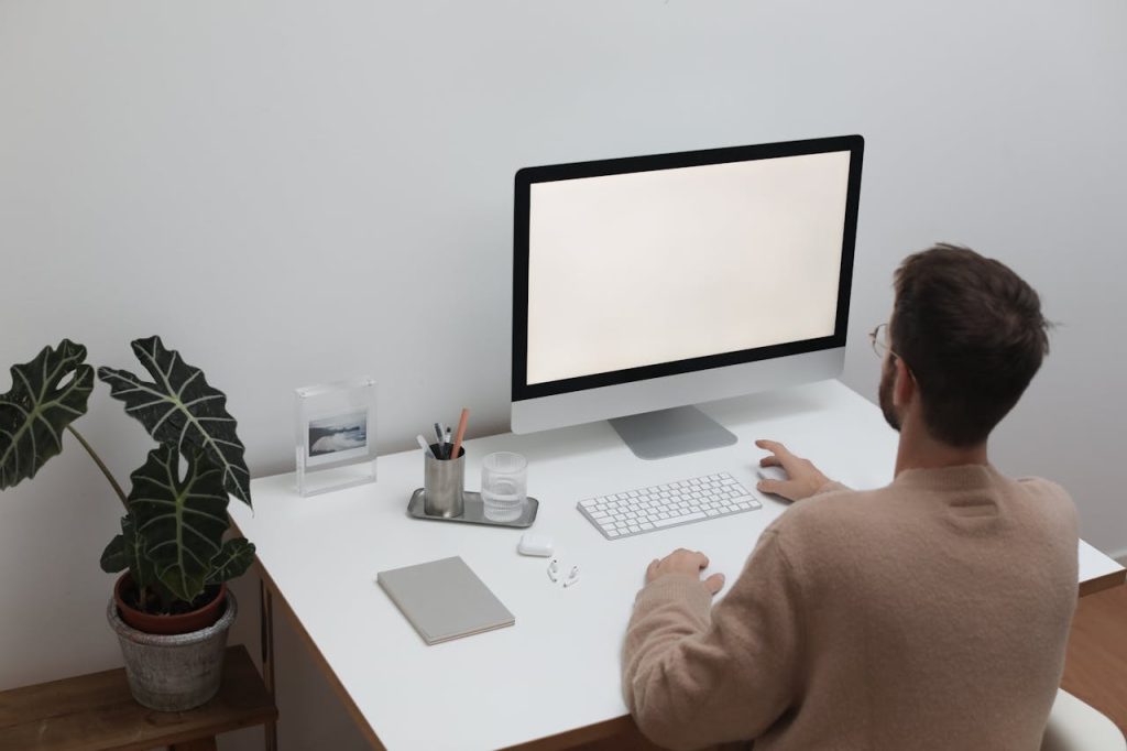 Back view of anonymous male in sweater sitting at table with notepad and earbuds while browsing computer