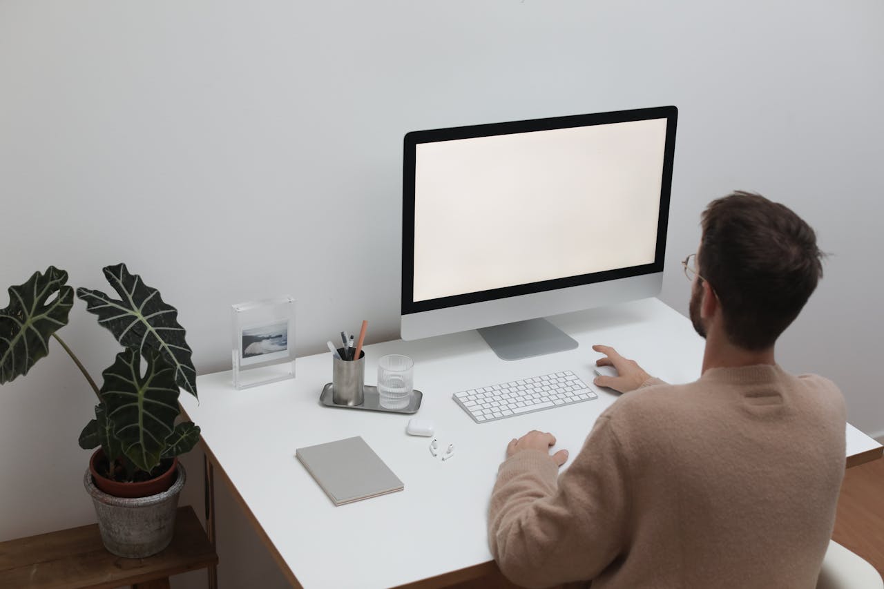 Back view of anonymous male in sweater sitting at table with notepad and earbuds while browsing computer